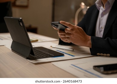 A man is using a cell phone while sitting at a desk. There are two cell phones on the desk, one of which is being used by the man