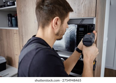 Man using cordless screwdriver to fix built in new microwave oven in modern kitchen cabinet. Concept of home appliance using