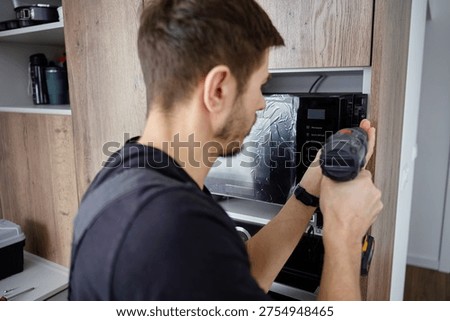 Man using cordless screwdriver to fix built in new microwave oven in modern kitchen cabinet. Concept of home appliance using
