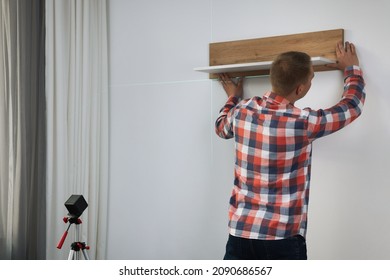 Man using cross line laser level for hanging wooden shelf on light wall indoors, back view