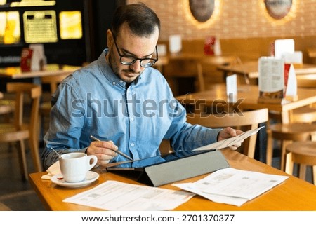 Man using a digital tablet with a stylus, organizing paper documents and financial statements while working from a restaurant