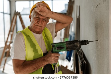 A man is using a drill at a construction site. He appears tired as he wipes his forehead. The area has tools and equipment. Bright light comes through the windows.