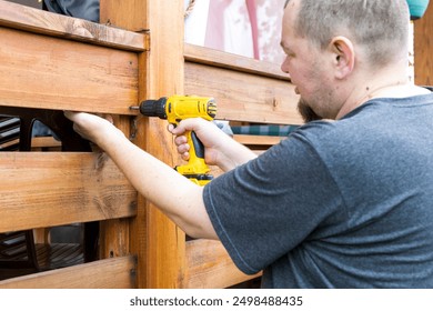 A man is using a drill to put a nail into a wooden fence. The man is wearing a gray shirt and has a beard