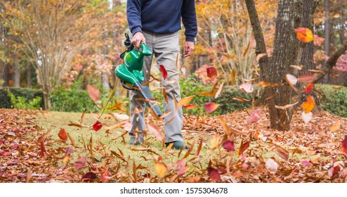 Man Using Electric Powered Leaf Blower To Blow Autumn Leaves From Grass Lawn. Landscape Worker Clearing Fall Leaves From Residential Yard.