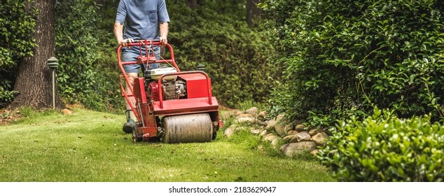 Man Using Gas Powered Aerating Machine To Aerate Residential Grass Yard. Groundskeeper Using Turf Aeration Equipment For Lawn Maintenance.