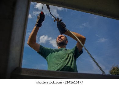 Man Is Using A Heat Torch Or Heat Gun To Apply Bitumen Insulation On The Edge Of A Roof Or Water Insulation On Concrete Edge Of A House Using A Ladder.