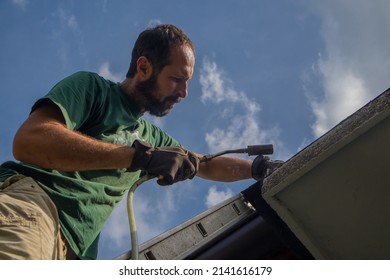 Man Is Using A Heat Torch Or Heat Gun To Apply Bitumen Insulation On The Edge Of A Roof Or Water Insulation On Concrete Edge Of A House Using A Ladder.