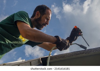 Man Is Using A Heat Torch Or Heat Gun To Apply Bitumen Insulation On The Edge Of A Roof Or Water Insulation On Concrete Edge Of A House Using A Ladder.