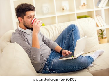 Man using laptop computer, sitting on sofa, drinking cup of coffee