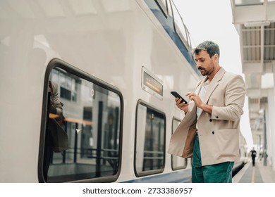 Man using a mobile phone, casually dressed, standing on a railway station platform next to a modern train, waiting to travel