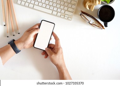 Man Using Mobile Phone At Office White Desk Table With Desktop Computer, Office Supplies And Coffee Mug. Top View Workspace And Blank Screen Smartphone