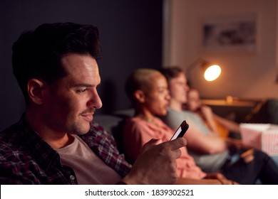 Man Using Mobile Phone Whilst Friends Watch TV At Home In Evening