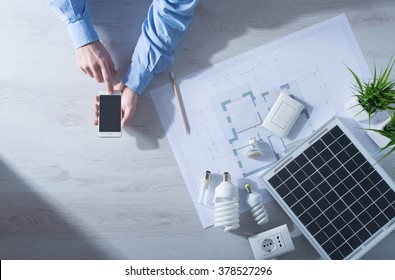 Man Using A Mobile Touch Screen Smart Phone Next To A Solar Panel, A Set Of CFL Lamps And A House Project, Energy Saving Concept