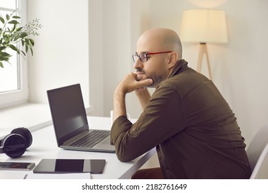 Man Using Multiple Digital Devices At Home. Pensive Bald Middle Aged Guy In Glasses And Casual Shirt Sitting At His Working Desk With Modern Laptop Computer, Tablet, Mobile Phone And Headphones