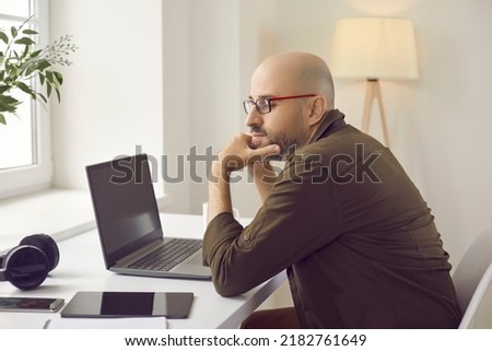 Man using multiple digital devices at home. Pensive bald middle aged guy in glasses and casual shirt sitting at his working desk with modern laptop computer, tablet, mobile phone and headphones