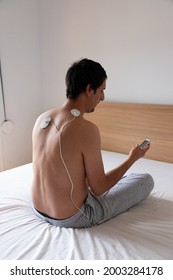 Man Using A Muscle Stimulator Machine On His Back And Neck Area Using Two Electrode Pads, Sitting On The Bed In Crossed Leg Position. White And Neutral Background.