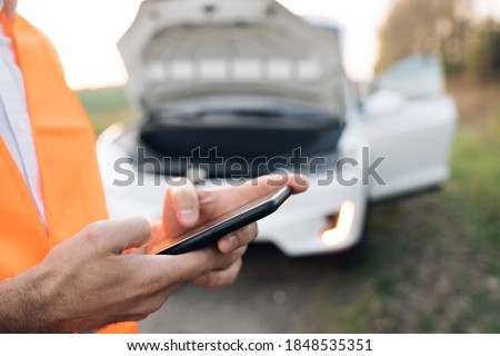 The man is using the phone in asking for help when his electric car is broken. Man in a safety vest. Concept road accident. Help repair.