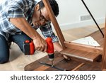 Man using a power drill to assemble wooden furniture. He is focused on securing a piece, wearing glasses and a plaid shirt in a home setting.