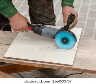 A man is using a power tool to cut a tile. The tile is white and the tool is blue