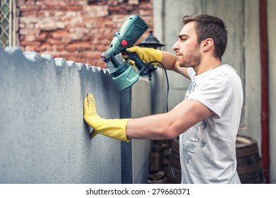 Man Using Protective Gloves Painting A Grey Wall With Spray Paint Gun. Young Worker Renovating House 