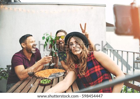 Man using rabbit ears gesture over head of friend in hat at table while eating pizza and drinking wine