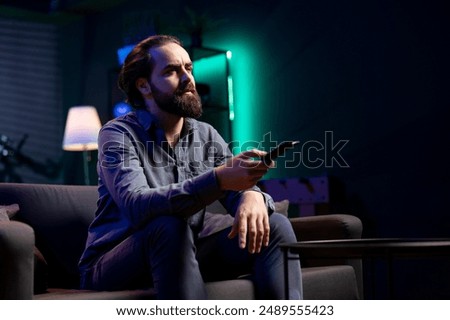 Man using remote control to switch channels, gripped by suspenseful films shown on television screen. Person mindlessly scrolling through streaming content, stopping after seeing interesting movie