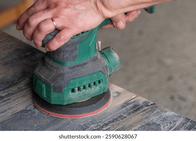 A man is using a sander to smooth out a surface. The sander is green and has a red handle. The man is wearing a ring on his finger
