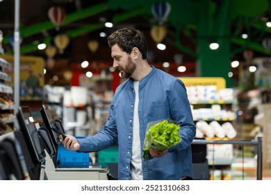 Man using self-checkout machine in busy supermarket environment holding fresh lettuce in one hand, phone in other. Image highlights modern shopping convenience, combining technology with grocery task
