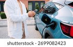 Man using smartphone app while standing next to his electric car charging at portable station on a city street, surrounded by urban buildings