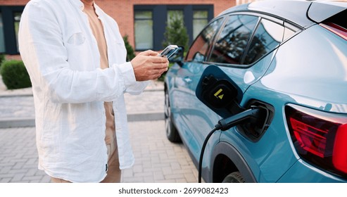 Man using smartphone app while standing next to his electric car charging at portable station on a city street, surrounded by urban buildings