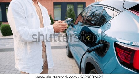 Man using smartphone app while standing next to his electric car charging at portable station on a city street, surrounded by urban buildings