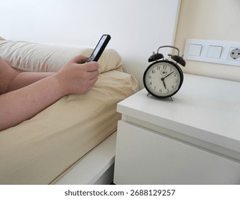 A man using a smartphone in bed next to a retro-style black alarm clock on a white bedside table. Hands of a fat woman with a smartphone in bed                   