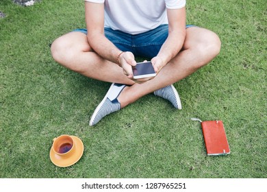 Man using smartphone and drinking coffee / tea on a grass.