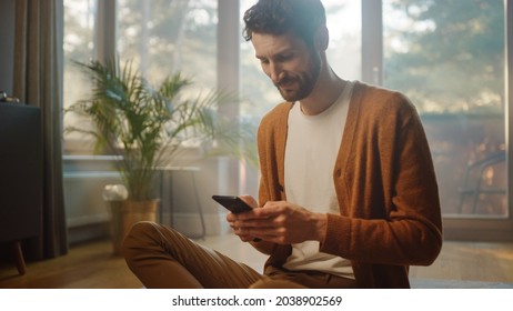 Man Using Smartphone at Home, Does Remote Work. Handsome Smiling Male Sitting on the Floor Works with Papers, Documents, Brainstorms Creative Project with Online Research. Sunny Living Room - Powered by Shutterstock