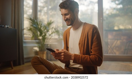 Man Using Smartphone at Home, Does Remote Work. Handsome Smiling Male Sitting on the Floor Works with Papers, Documents, Brainstorms Creative Project with Online Research. Sunny Living Room - Powered by Shutterstock