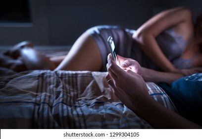 Man Using Smartphone And Ignoring Sexy Woman In Bed At Night. Looking At Mobile Phone. Husband Giving No Attention To Wife. Disregarding Marriage. Couple Problem. Cellphone Addiction. Obsessed Addict.