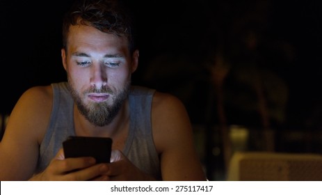 Man Using Smartphone At Night Browsing Internet Updating Social Media. Young Male With Beard Using Mobile Cell Smart Phone Outdoors In The Dark In Summer.