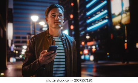 Man Using Smartphone Walking Through Night City Street Full of Neon Light. Smiling Stylish Man Using Mobile Phone, Social Media, Online Shopping, Texting on Dating App. - Powered by Shutterstock