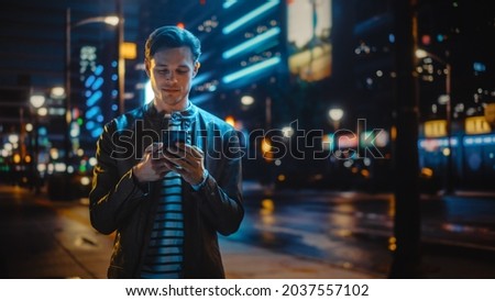 Man Using Smartphone Walking Through Night City Street Full of Neon Light. Smiling Stylish Man Using Mobile Phone, Social Media, Online Shopping, Texting on Dating App.