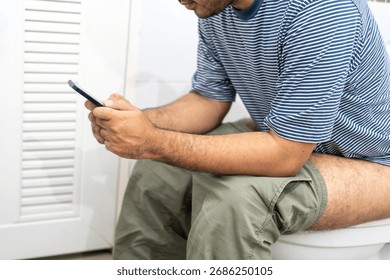 Man using smartphone while sitting on toilet bowl in bathroom at home.