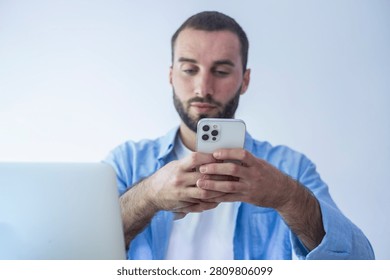 Man using smartphone while working on laptop at home, focused on his phone screen, remote work, multitasking and communication in a modern, casual office or home workspace