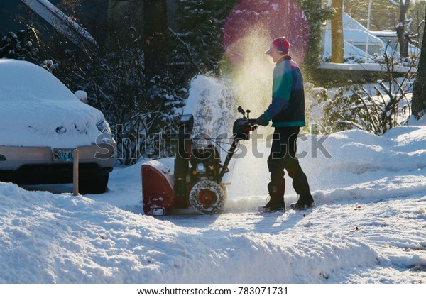 A man using a snow blower to clear snow from his\
driveway as the sun sets and reflects light off the snow to produce\
colorful lens flares. A juxtaposition between cold temps and warm\
sun rays.