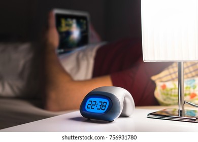 Man Using Tablet Or Watching Movie Or Series Stream Online In Bed Before Falling Asleep. Digital Alarm Clock On Nightstand In Bedroom.