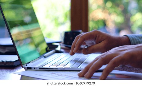 Man using touch screen laptop at desk, backlit by sunshine with reflections through window, close up.