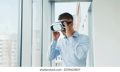 Man Using Virtual Reality Headset in Modern Office. A young man in a light blue shirt explores virtual reality using a VR headset in a bright, modern office with large windows.