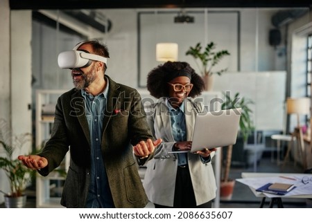 Man using virtual reality simulator headset while his african female coworker standing beside him and holding laptop