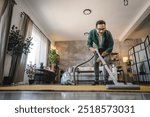 A man is vacuum the floor with vacuum cleaner in a stylish, contemporary living room filled with plants and soft lighting, reflecting a tidy and organized home environment