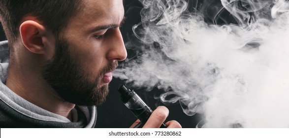 Man with vaping mod exhaling steam at black studio background. Bearded guy smoking e-cigarette to quit tobacco. Vapor and alternative nicotine free smoking concept, closeup