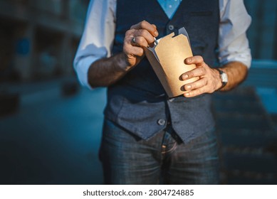 Man in a vest and jeans opening a rectangular cardboard food box, taking a quick lunch break or snack outdoors in an urban environment during daylight