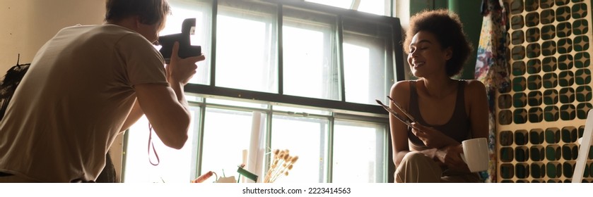 Man With Vintage Camera Taking Photo Of Smiling African American Girlfriend With Cup And Paintbrushes, Banner